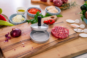 Burger press on wooden cutting board, vegetables and spices with kitchen bowls.