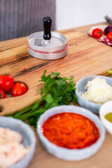 Burger press on wooden cutting board, vegetables and spices with kitchen bowls.