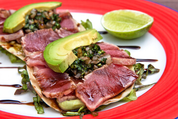 Mexican tostadas with shaved tuna, onion seasoned with soy sauce, slices of avocado and lemon, served on a red and white plate on a wooden background