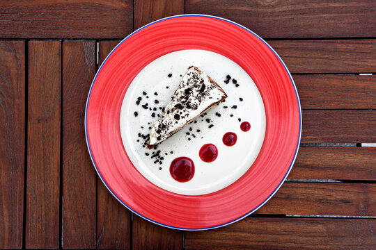 Slice of cheesecake covered in whipped cream and chocolate cookie, served on a red plate with white on wooden background, top view
