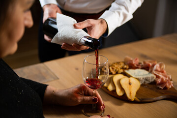 A man in a white shirt pouring guests a red wine to a wine glass from a bottle. Wine tasting with a cheese board. 