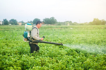 Obraz premium A farmer with a mist sprayer blower processes the potato plantation from pests and fungus infection. Protection and care. Fumigator fogger. Use of agriculture industrial chemicals to protect crops.