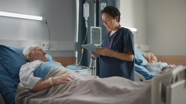 Hospital Ward: Friendly Head Nurse Talks with Elderly Patient Resting in Bed. Physician Uses Tablet Computer, Does Checkup, Ask Health Care Questions. Old Man Fully Recovering after Successful Surgery