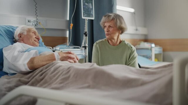 Hospital Ward: Elderly Man Resting in Bed, His Caring Beautiful Wife Supports Him Sitting Beside, Happy together They Talk about Good Times. Old Man Fully Recovering after Successful Surgery