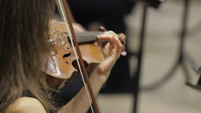 Cinematic close-up shot of female violinist is playing violins during musical concert on classic theatre stage with symphony orchestra performing on background.