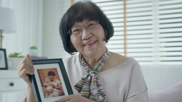 Portrait of happy senior asian woman relax sitting smile looking at photo frame reminding the past memory at home with enjoy retirement or quarantine time. Asia adult older people happy lifestyle.