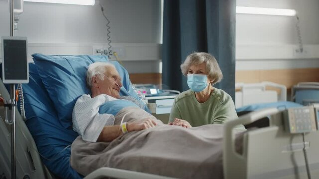 Hospital Ward: Elderly Man Resting in Bed, Caring Beautiful Wife Wearing Face Mask Visits Beloved Husband, Supports Him Sitting Beside, Holding Hands, Happy Couple Talking. Recovering After Surgery