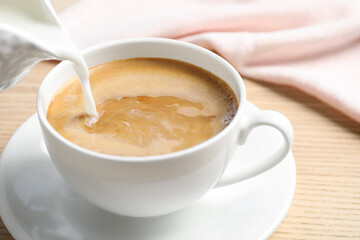 Pouring milk into cup of hot coffee on wooden table, closeup