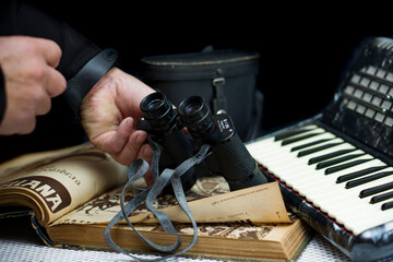 hands holding vintage binoculars over an antique book and an accordion - obrazy, fototapety, plakaty