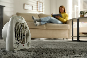 Woman reading book in living room, focus on electric fan heater - obrazy, fototapety, plakaty