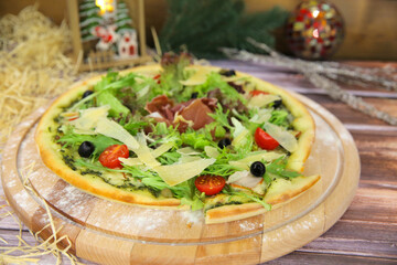 Side view of Italian pizza served on wooden plate with Christmas new year decoration on background. Fast food dish with fresh lettuce, jamon Serrano ham slices, Parmesan cheese, olives, tomatoes
