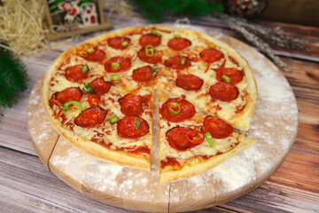 Close up of Cheesy Italian pepperoni pizza served on wooden plate and table with Christmas new year decoration on background. Fast food dish with salami sausages, red and green chili pepper slices
