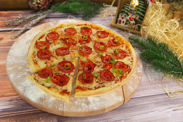 Side view of cheesy Italian pepperoni pizza served on wooden plate and table with Christmas new year decoration on background. Fast food dish with salami sausages, red and green chili pepper slices
