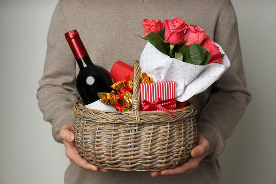 Man holding wicker basket with gifts, bouquet and wine on grey background, closeup