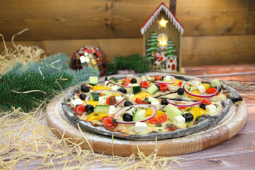 Side view Italian black dough veggie pizza served on wooden plate and table with Christmas new year decoration on background. Fast food vegetable dish with fresh cucumber, tomato, paprika, feta cheese