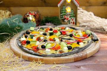 Italian black dough vegetarian pizza served on wooden plate and table with Christmas new year decoration on background. Fast food vegetable dish with fresh cucumber, tomato, paprika, feta cheese
