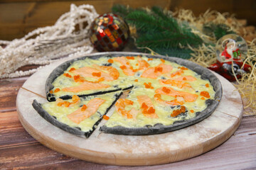 Side view of Italian black dough pizza served on wooden plate and table with Christmas decoration on background. Fast food seafood dish with red fish salmon slices, black tiger shrimps and red caviar
