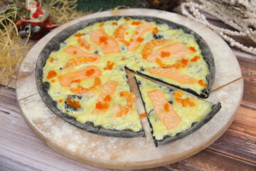 Italian black dough pizza served on wooden plate and table with Christmas new year decoration on background. Fast food seafood dish with red fish salmon slices, black tiger shrimps and red caviar
