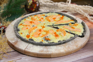 Close up of Italian black dough pizza served on wooden plate and table with Christmas new year decoration on background. seafood dish with red fish salmon slices, black tiger shrimps and red caviar
