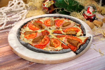 Italian black dough pizza served on wooden plate and table with Christmas new year decoration on background. Fast food meat dish with pork, beef, tomato slices and mozzarella cheese 
