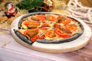 Foto op Plexiglas Side view of Italian black dough pizza served on wooden plate and table with Christmas new year decoration on background. Fast food meat dish with pork, beef, tomato slices and mozzarella cheese   © Art Food