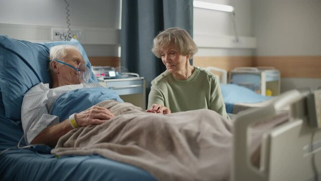 Hospital Ward: Elderly Man Wearing Oxygen Mask Resting in Bed, Caring Beautiful Wife Visits Beloved Husband, Supports Him Sitting Beside, Holding Hands, Happy Couple Talking. Recovering After Surgery