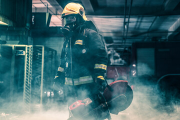 Portrait of a firefighter wearing full protective equipment posing with a chainsaw on his shoulder. Dark background with smoke and blue light. - obrazy, fototapety, plakaty