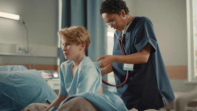 Hospital Ward: Handsome Young Boy Resting in Bed with Caring Mother Supporting Him, Friendly Head Nurse Listens to His Lungs, Heart Beat with Stethoscope
