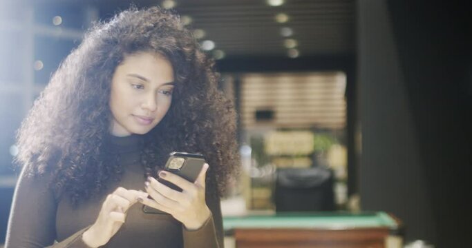 Close-up of attractive Latin American girl with curly hair and perfect healthy skin holding mobile phone and looking at screen with concentrated expression while watching videos online. 4K