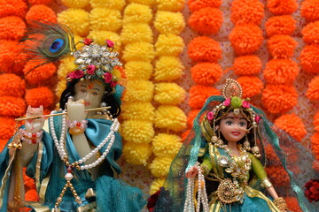 Idol of Krishna and Radha beautifully dressed and decorated in colorful attire and jewelry, with a background of orange and yellow flowers, shot in landscape composition