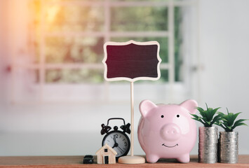 Pink piggy bank, coin money, alarm clock, house model, Toy cars, and signs, all placed on the table In a white office., the concept of saving money for the future, space for text.