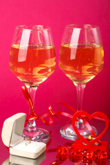 Two glasses of champagne tied with red ribbons on a pink background next to a ring in a box and a candle in a candlestick.