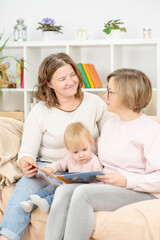 Two young women are sitting in the living room on the couch with a child on their knees and are reading a book. Gay lesbian couple at home with a baby. Lesbian family concept