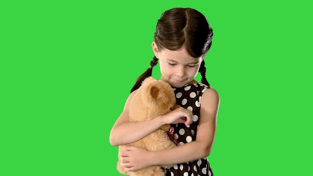 Shy little girl in polka dot dress hugging her teddy bear while walking and looking down at her feet on a Green Screen, Chroma Key.