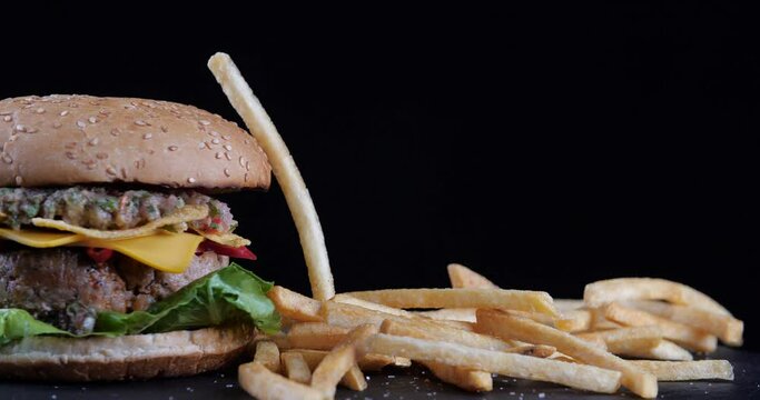 Cropped shot of large hearty appetizing hamburger bun with cheese salad leaves beef tomatoes and grilled sauce stands on black table, yellow crispy fries falling on top of surface, fast food concept