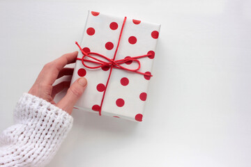 Female hand in white sweater holding a gift box in red polka dot pattern wrapping paper on a white background. Top view, copy space 
