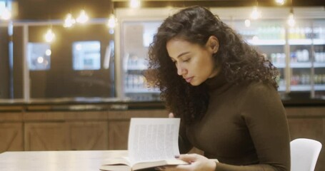 Concentrated Latin female student in library with modern interior and reading literature. intelligent woman interested on book for leaders. Curly hair Brazilian girl. 4K.