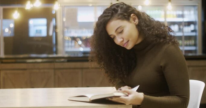 Concentrated Latin female student in library with modern interior and reading literature. intelligent woman interested on book for leaders. Curly hair Brazilian girl. 4K.
