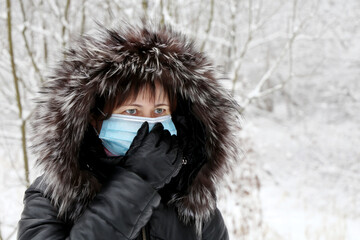 Coronavirus protection, woman in medical mask and fur hood standing in winter park during snow. Concept of illness, fever, cold and flu