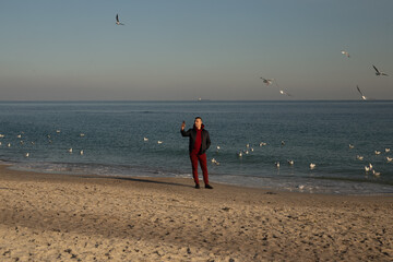 A guy in a red suit and jacket makes a selfie in the winter near the sea. Seagulls are flying over the sea, against the background of a guy in a red suit.