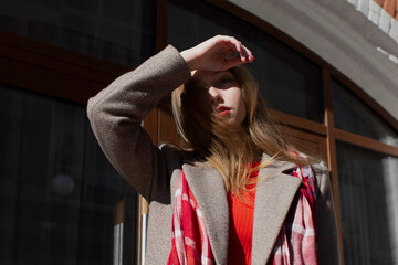 Portrait of a young woman in coat covering face by hand of bright sunlight standing on street in front of doors. Cheerful girl having fun outside. Urban style and fashion. Casual outfit