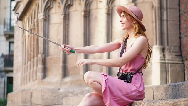 Cute woman taking a selfie with a stick and a phone. Girl in sweet pink dress and a hat using her smartphone to take pictures of herself by a church in Barcelona.