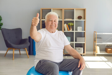 Happy senior man giving thumbs-up and looking at camera sitting on yoga ball at home