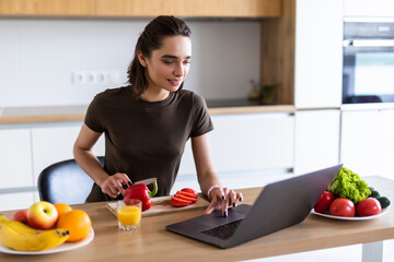 Beautiful woman cutting bell pepper while watch laptop in the kitchen