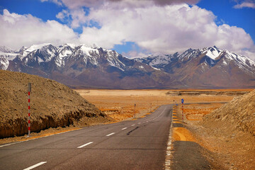 Highway stretching across the brown Himalaya Mountains, against a blue sky covered by white clouds.