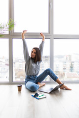 Portrait of a cheerful woman sitting on the floor with computer and raised hands up at home