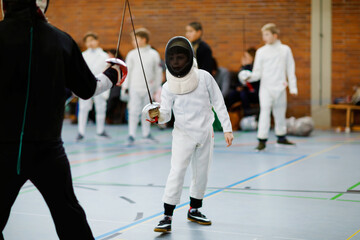 Little kid boy fencing on a fence competition. Child in white fencer uniform with mask and sabre. Active kid training with teacher and children. Healthy sports and leisure.