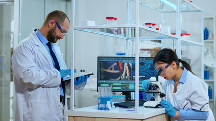 Team of scientists doing biological researches under microscope, lab technician writing data into tablet in modern equipped laboratory. Multiethnic group examining virus evolution using high tech.