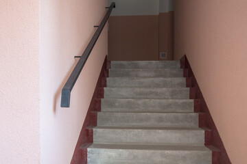 Gray concrete staircase with maroon stripes on the sides inside the entrance with walls painted in peach color with a black metal handrail mounted in the wall in apartment building.