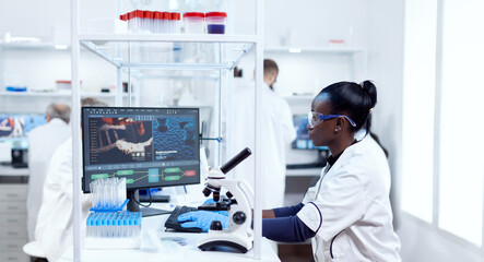 African scientist working on computer in modern facility and colleagues in the background. Black...